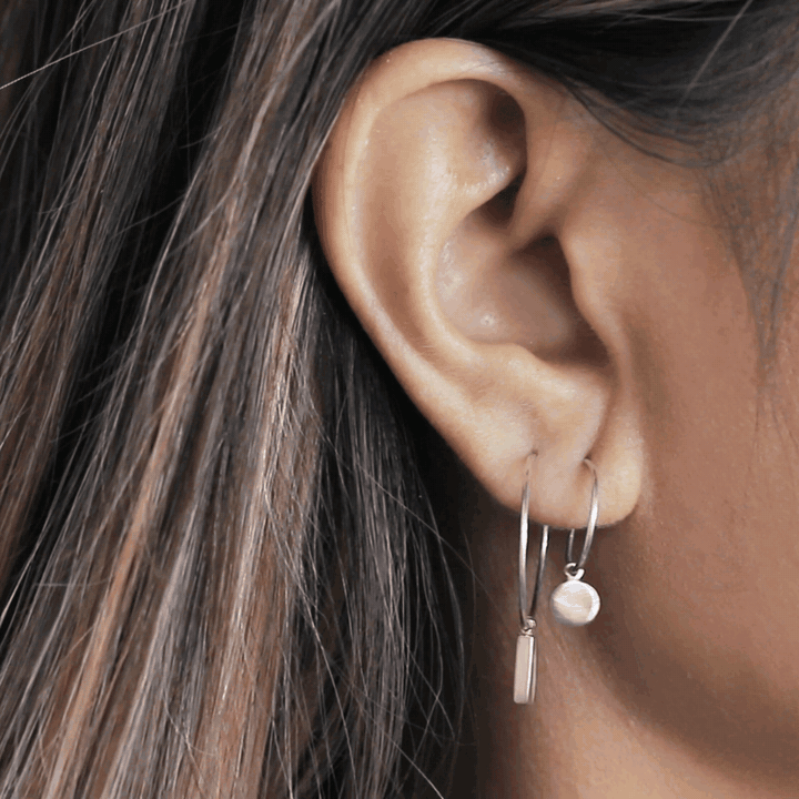 An animation of two silver hoop earrings, one with a bar charm and one with a small disc, worn on an ear.