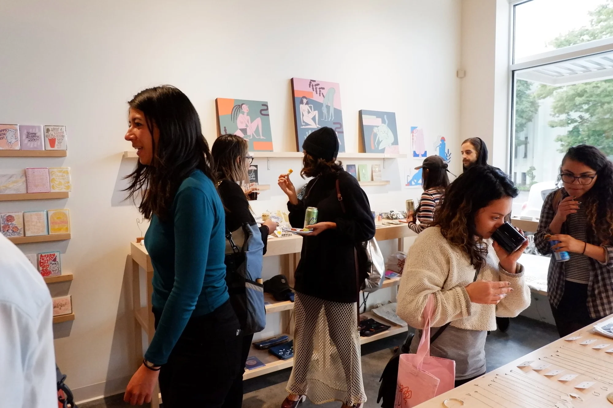 Visitors holding drinks and browsing cards, paintings and jewelry in the sunlit shop.