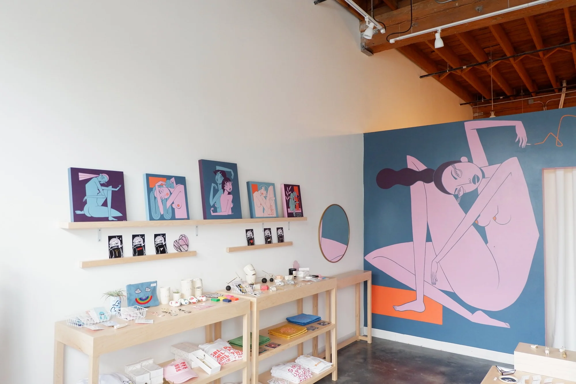 Paintings and small prints on shelves beside a table of apparel and goods, with the mural wall behind.