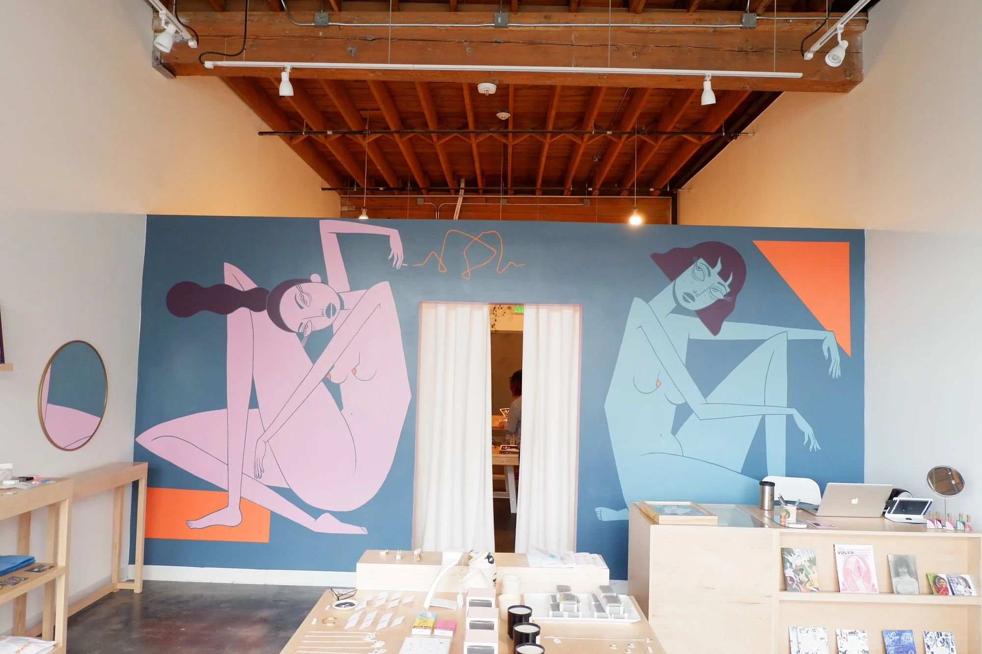 The inside of Project Object, a bright shop with a large blue, pink and orange mural wall, a white curtain doorway and tables of jewelry and candles.