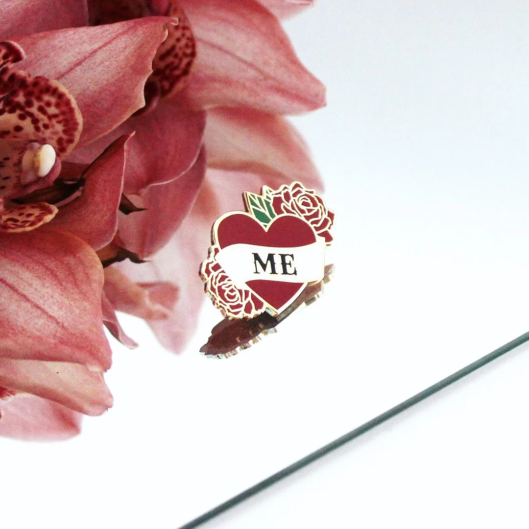An enamel pin shaped like a red heart with the word ME on a banner and roses, photographed on a mirror beside pink orchids.
