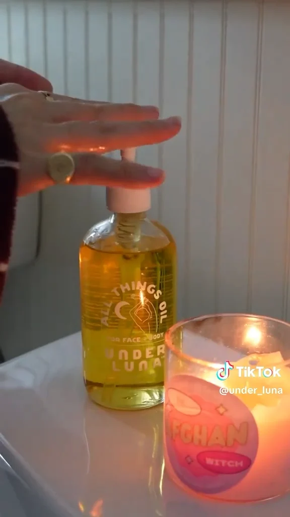 A frame from the Under Luna TikTok: a hand pressing the pump on a bottle of All Things Oil, next to a lit candle.