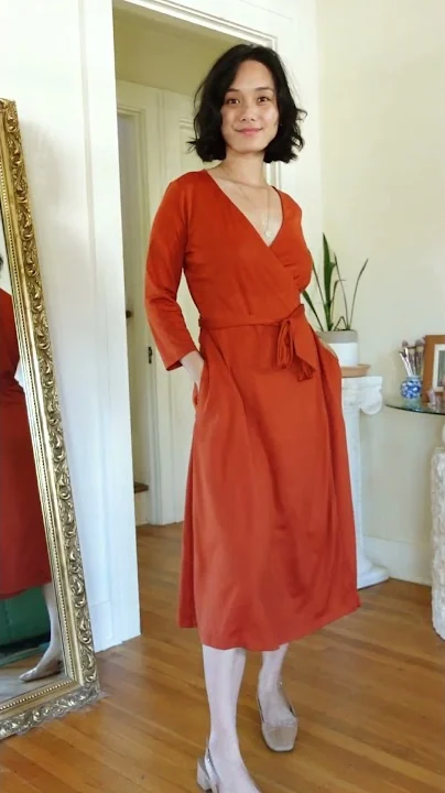 A frame from the wool& Short: Margaret in a rust-colored merino wrap dress, standing in a bright room beside a gold mirror.